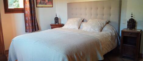 A bedroom with a large bed, a headboard, two nightstands, a window with curtains, and a wall-mounted artwork.