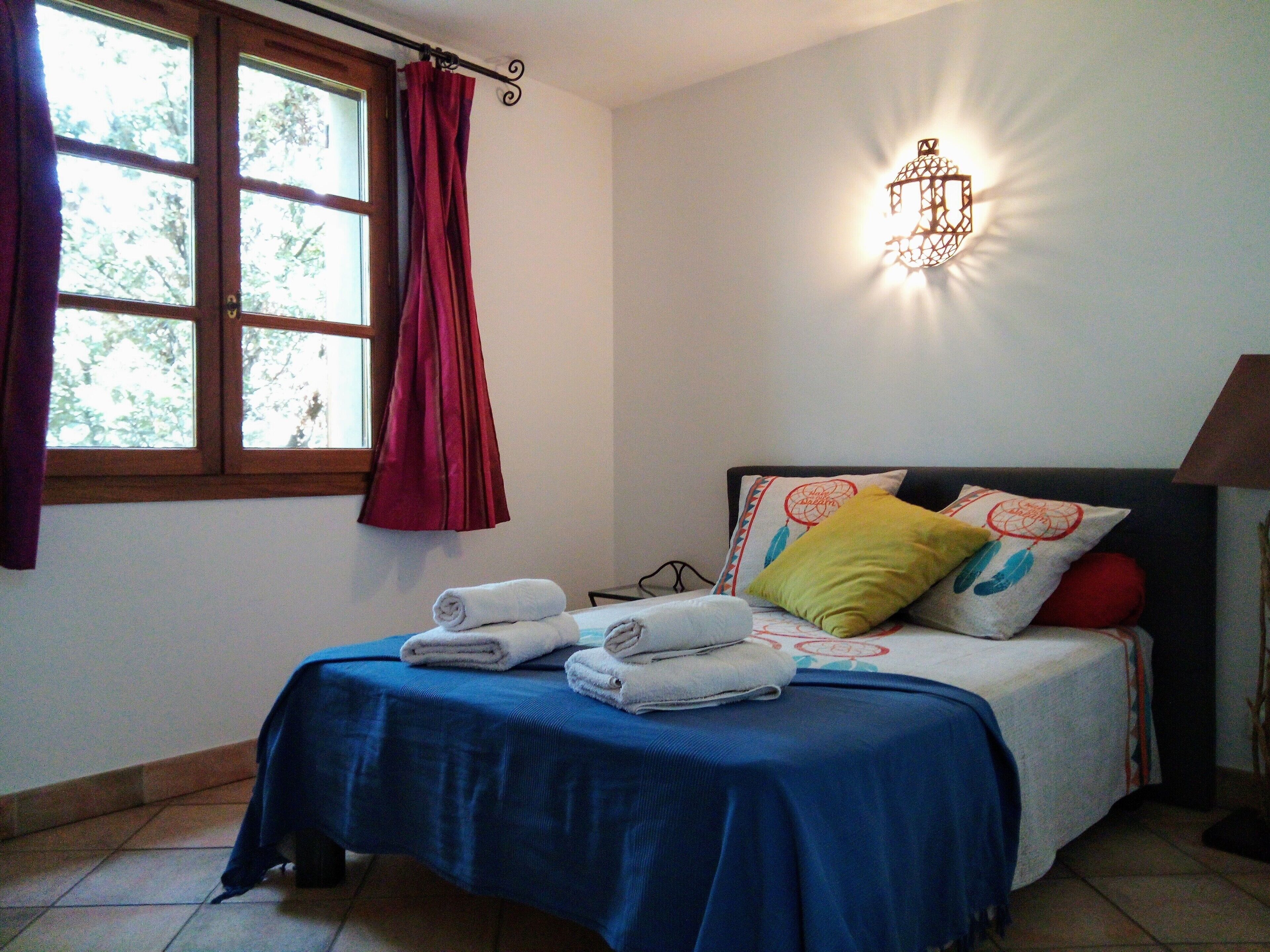 A bedroom with a bed, a window with curtains, a wall-mounted light fixture, and a bedside table.