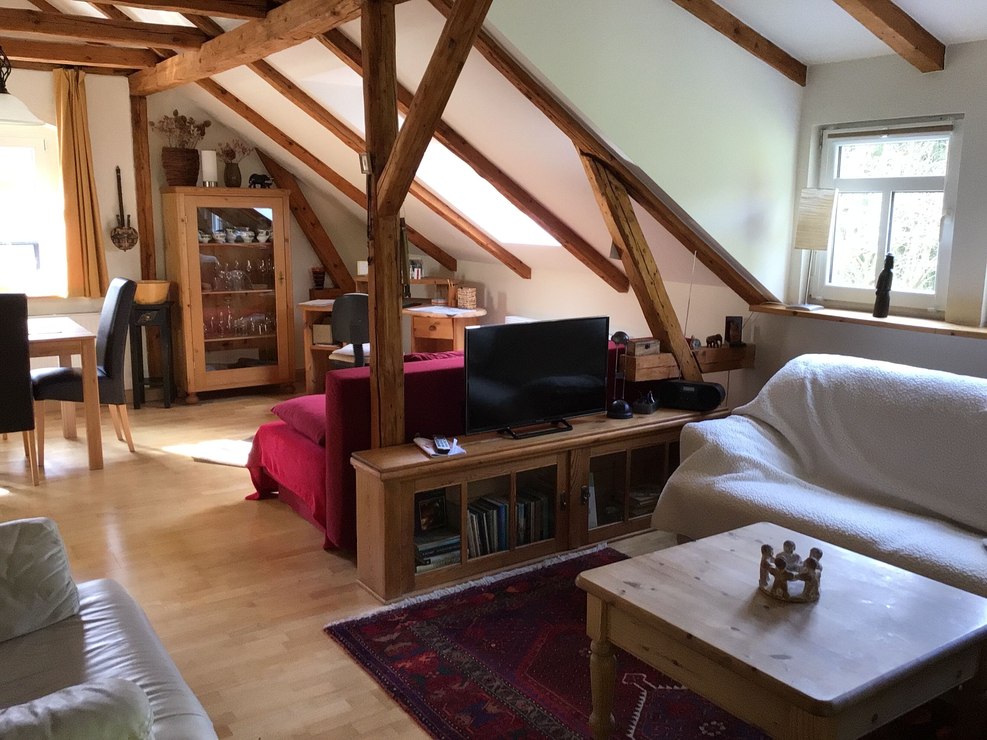 Un habitación estilo loft con vigas de madera expuestas, un televisor sobre un soporte de madera, un sofá rojo, un sofá blanco y una pequeña mesa con una figurita.