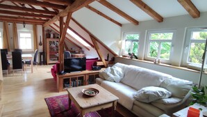 A cozy loft-style living room with exposed wooden beams, a stone fireplace, a dining area with a table and chairs, and large windows offering a view of greenery. - Stylishly furnished apartment in Rathmannsdorf in the Zauketal, allergy sufferers (Rathmannsdorf)