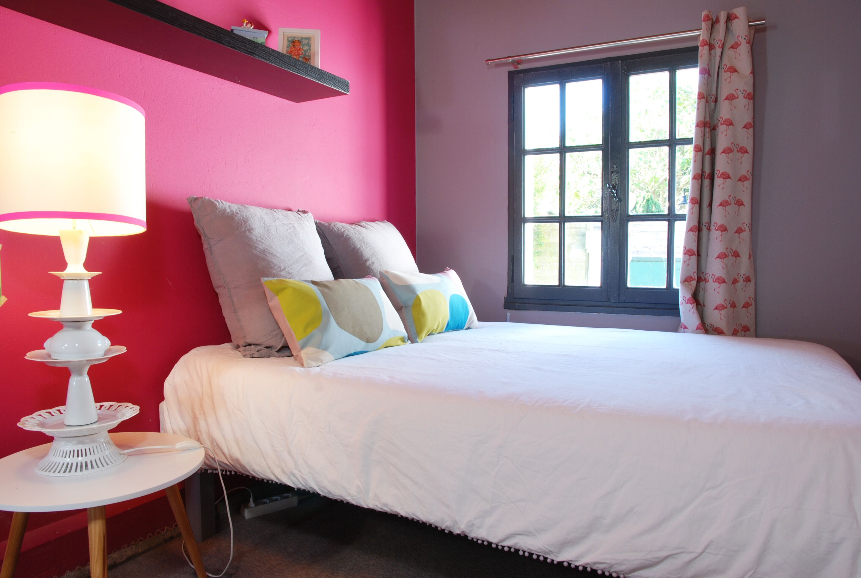 A bedroom with a bed, a bedside table with a lamp, a window with curtains, and a pink wall.