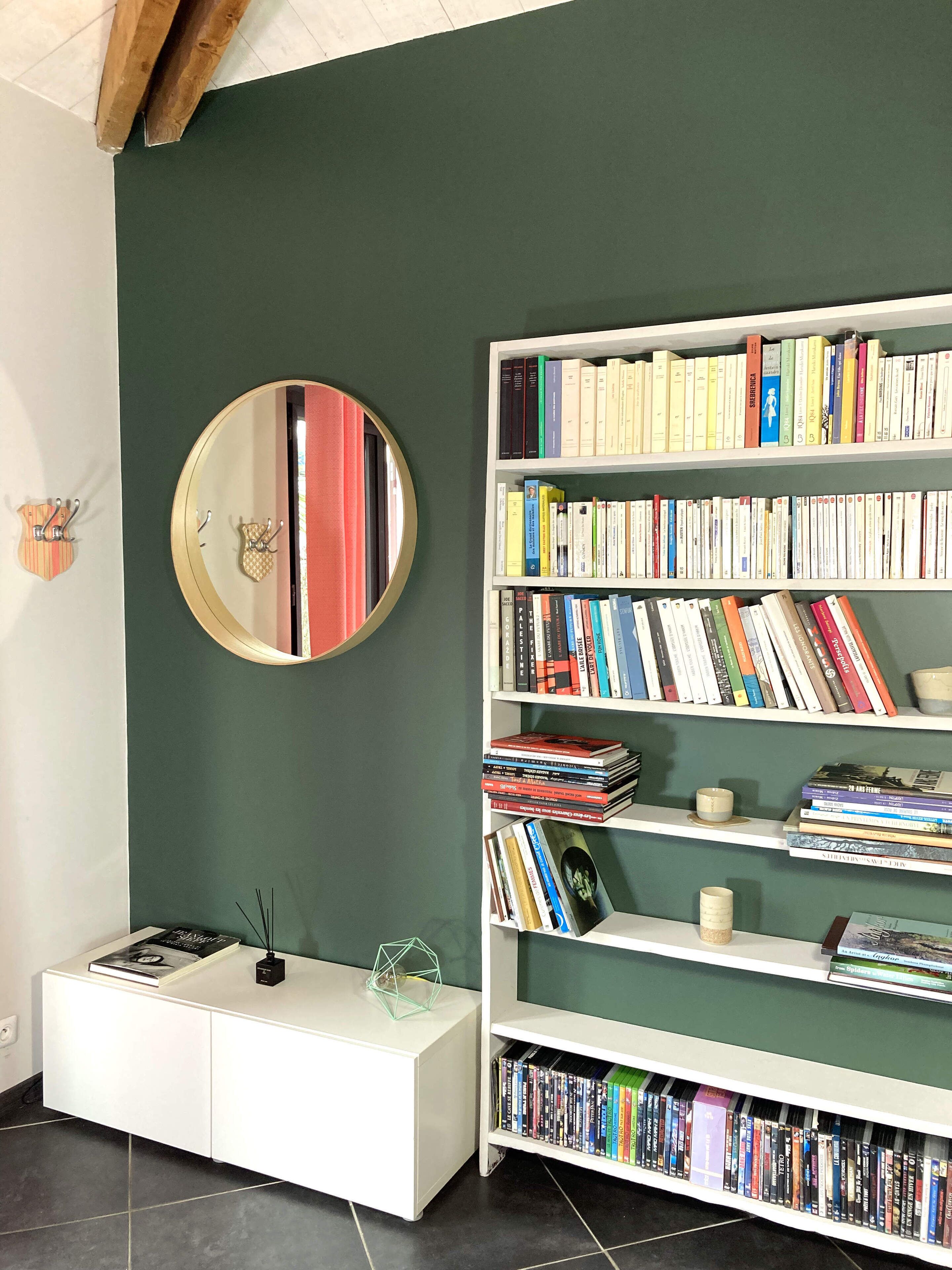 A room with a bookshelf filled with books, a round mirror on the wall, and a white side table with a book and a candle.