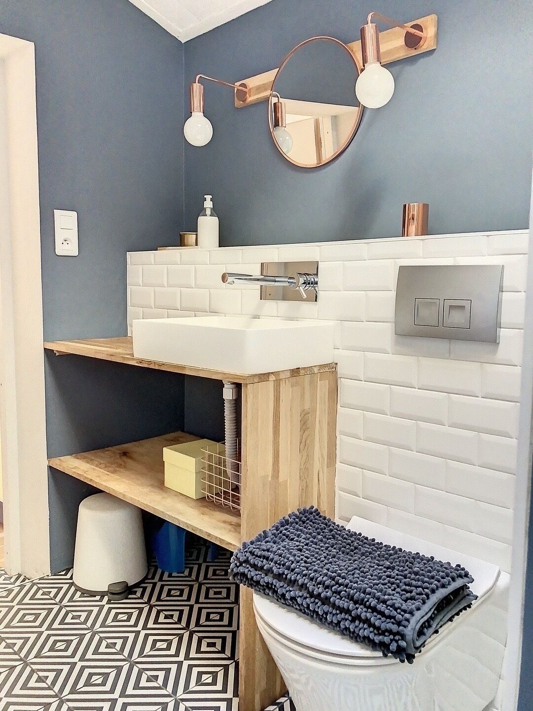 Een moderne badkamer met een witte wasbak, houten planken, een ronde spiegel en een vloer met een patroon.