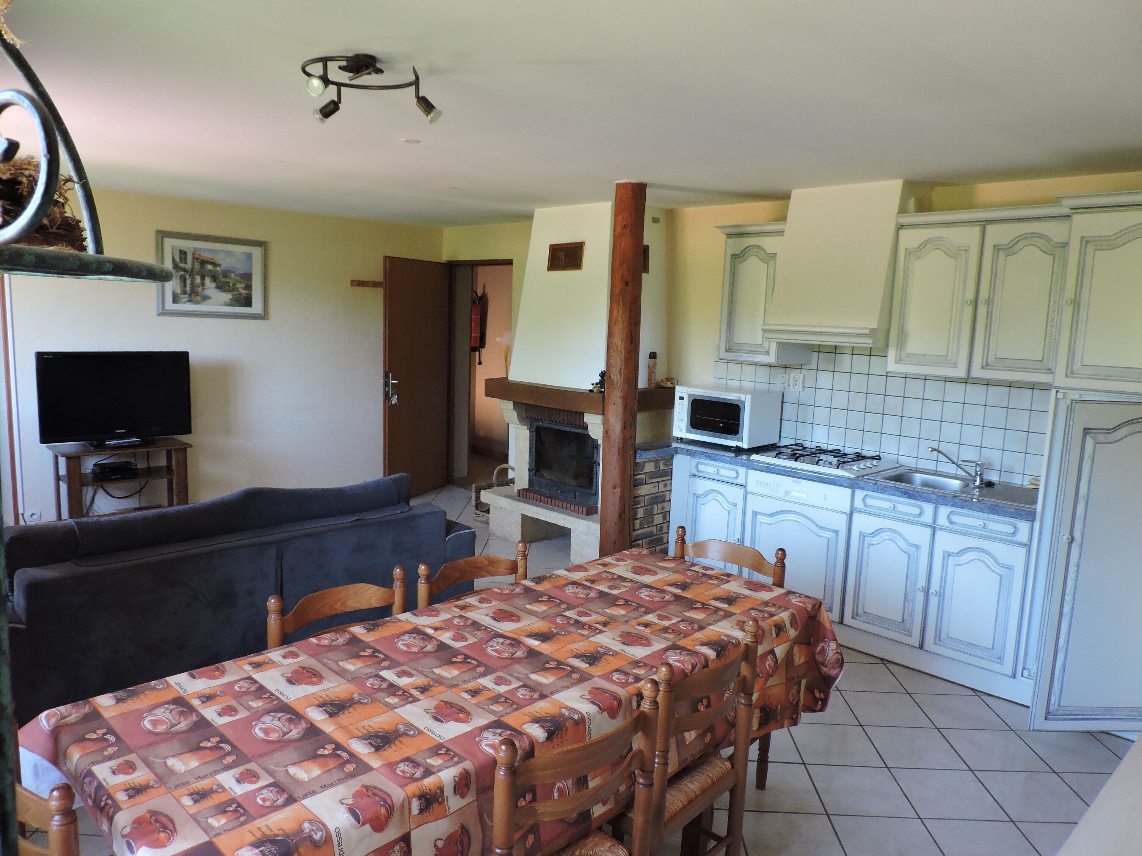 Un comedor con una mesa lista para cuatro, televisor, chimenea y una cocina con armarios blancos y microondas.