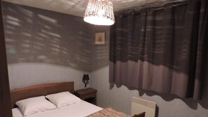 A bedroom with a wooden bed, a nightstand with a lamp, a radiator, and a window with curtains. - Rental cottage in renovated farm (Arrentès-De-Corcieux)