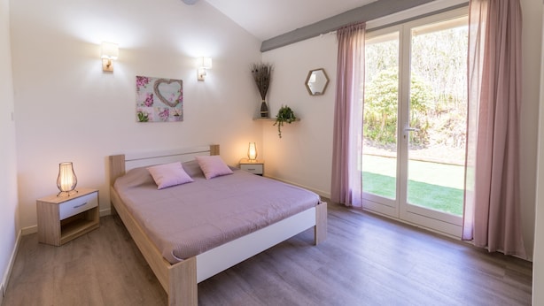 Une chambre à coucher comprenant un lit, des tables de chevet, des lampes fixées au mur, un élément décoratif mural et une vue sur un jardin, accessible par une porte coulissante.