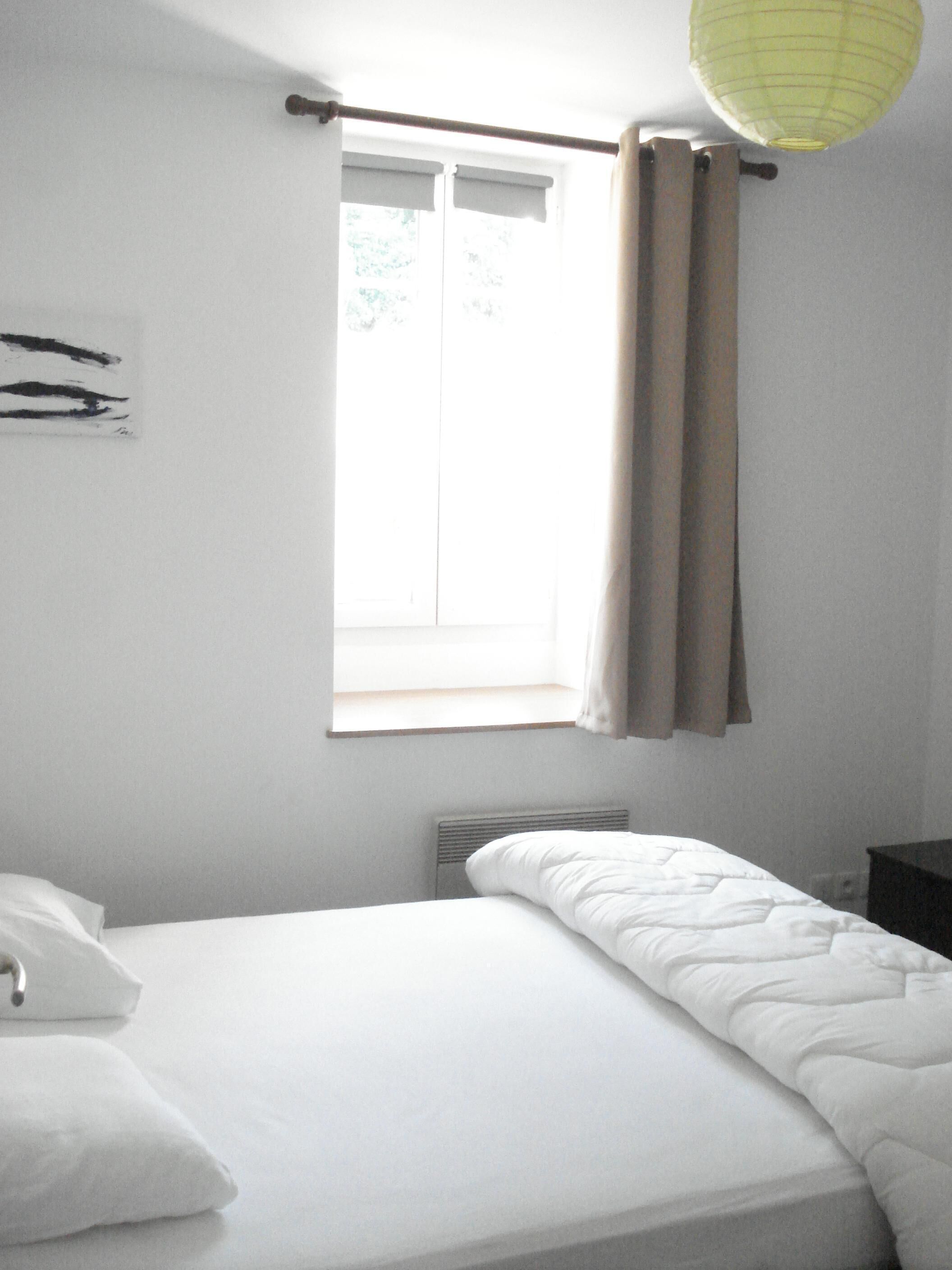 Un dormitorio con cama, ventana con cortinas, radiador y una lámpara fijada a la pared.