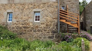 Un bâtiment en pierre avec un escalier en bois menant à une fenêtre, entouré d’un jardin où poussent diverses plantes et fleurs.