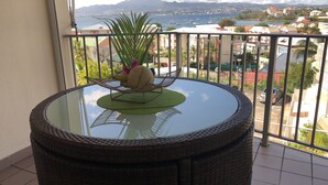 A round glass table with a wicker base, a small green mat, and a decorative item on a balcony overlooking a coastal town with buildings and boats. - RESIDENCE VERY CALM  (Anse mitan)