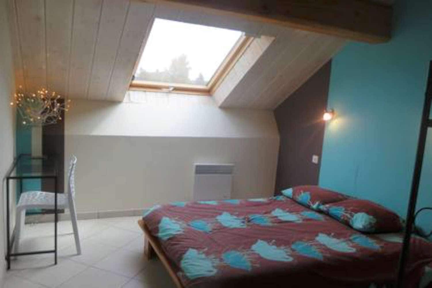 A bedroom with a sloped ceiling, a skylight, a bed with patterned bedding, a chair, and a bedside table.