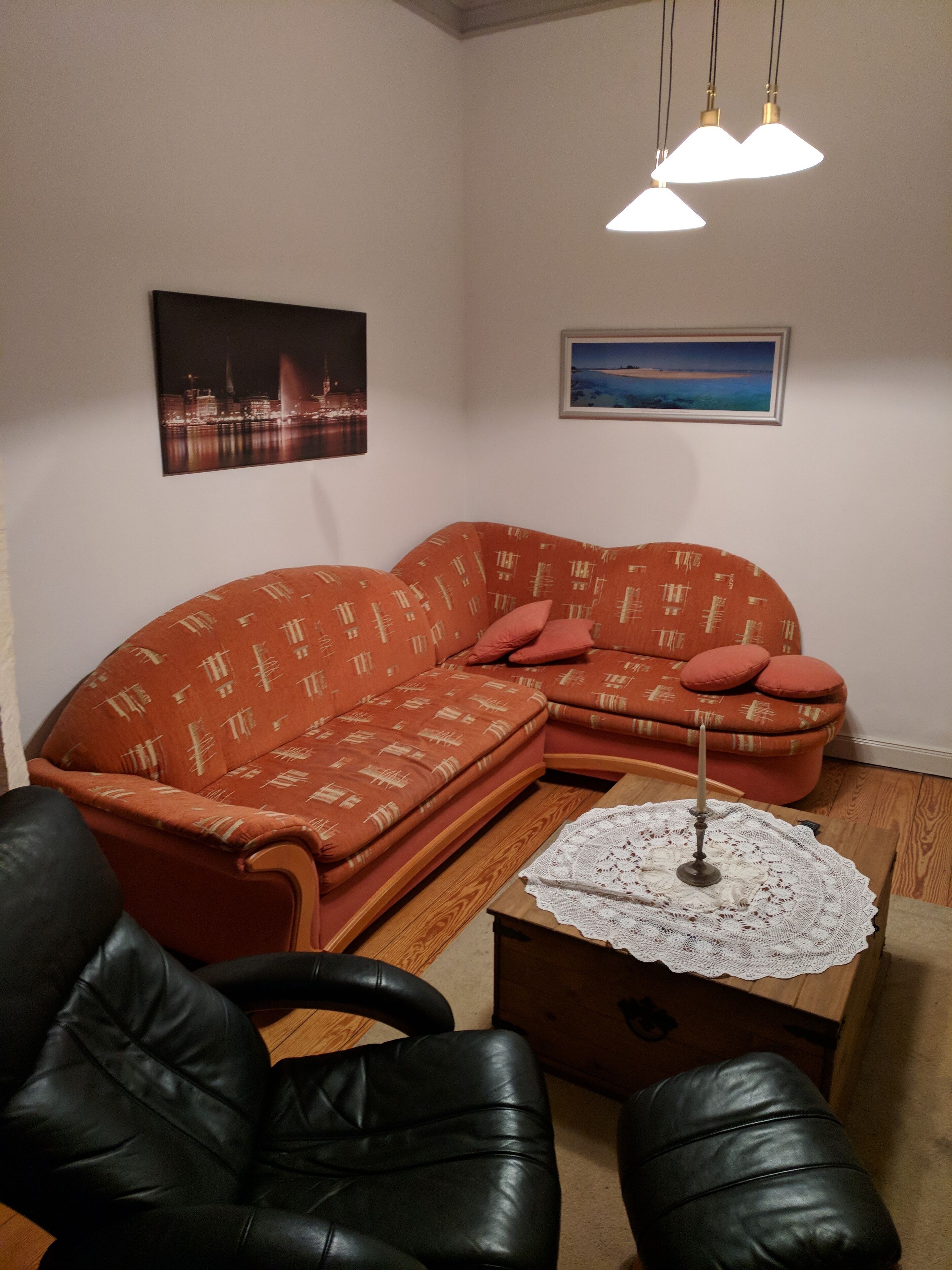 Een woonkamer met een hoekbank, een zwarte leren fauteuil, een houten salontafel met een kantkleed en twee hanglampen.