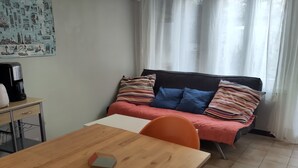 Ein kleiner, gemütlicher Raum mit Holztisch, Bett mit bunten Kissen, einer Kaffeemaschine und einem Wandposter.