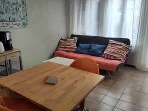 Een knus kamertje met een houten tafel, een bed met kleurrijke kussens, een koffiezetapparaat en een poster aan de muur.