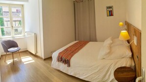 Un dormitorio con una cama, una silla, una ventana, una lámpara y un cuadro en la pared.