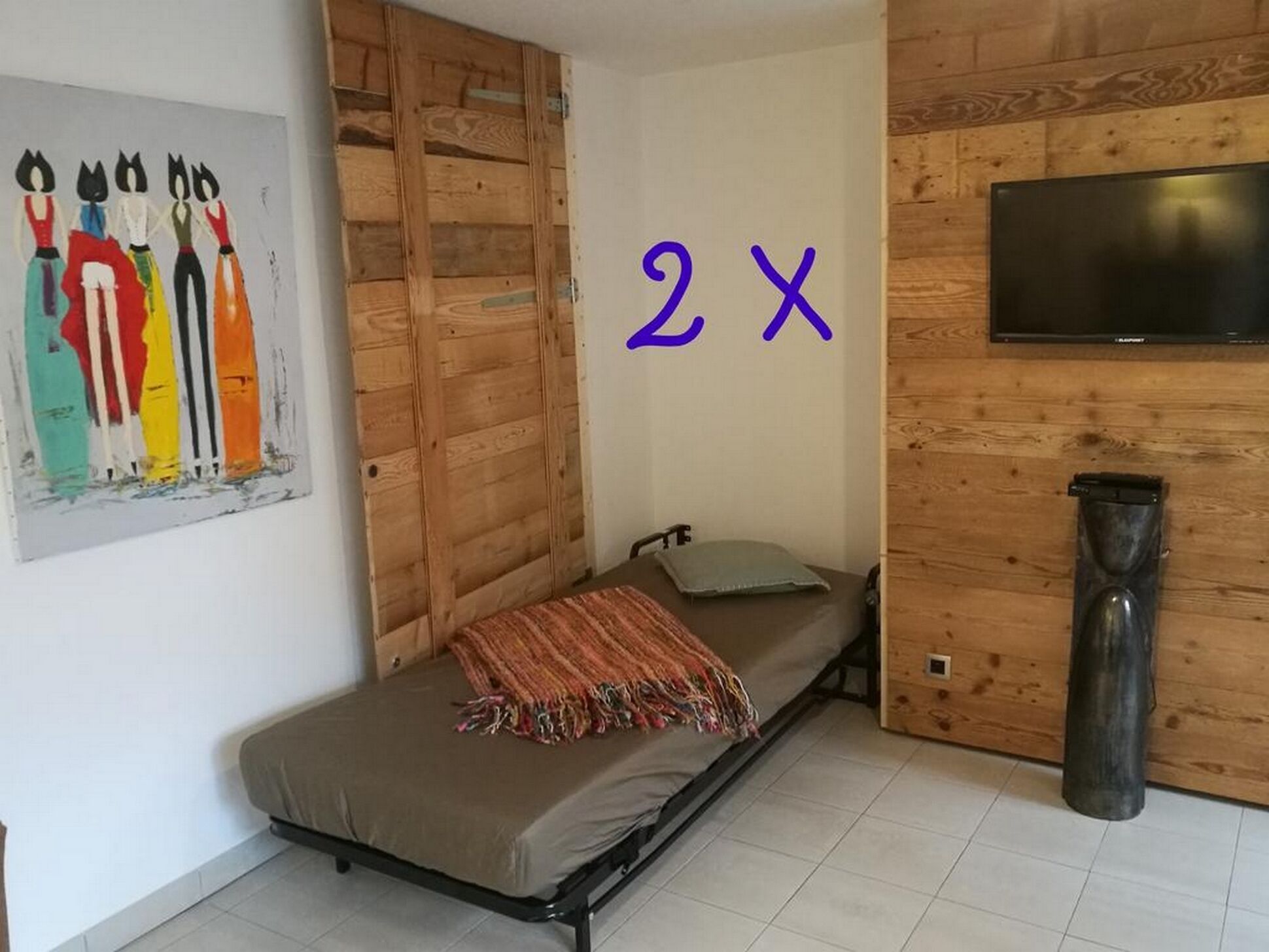 Une petite pièce avec un lit simple, un mur en bois, un téléviseur fixé au mur et un tableau accroché au mur.