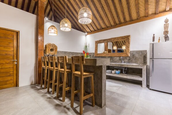 Une cuisine avec des chaises en bois, un comptoir de bar, des luminaires suspendus, un réfrigérateur et un grand miroir.