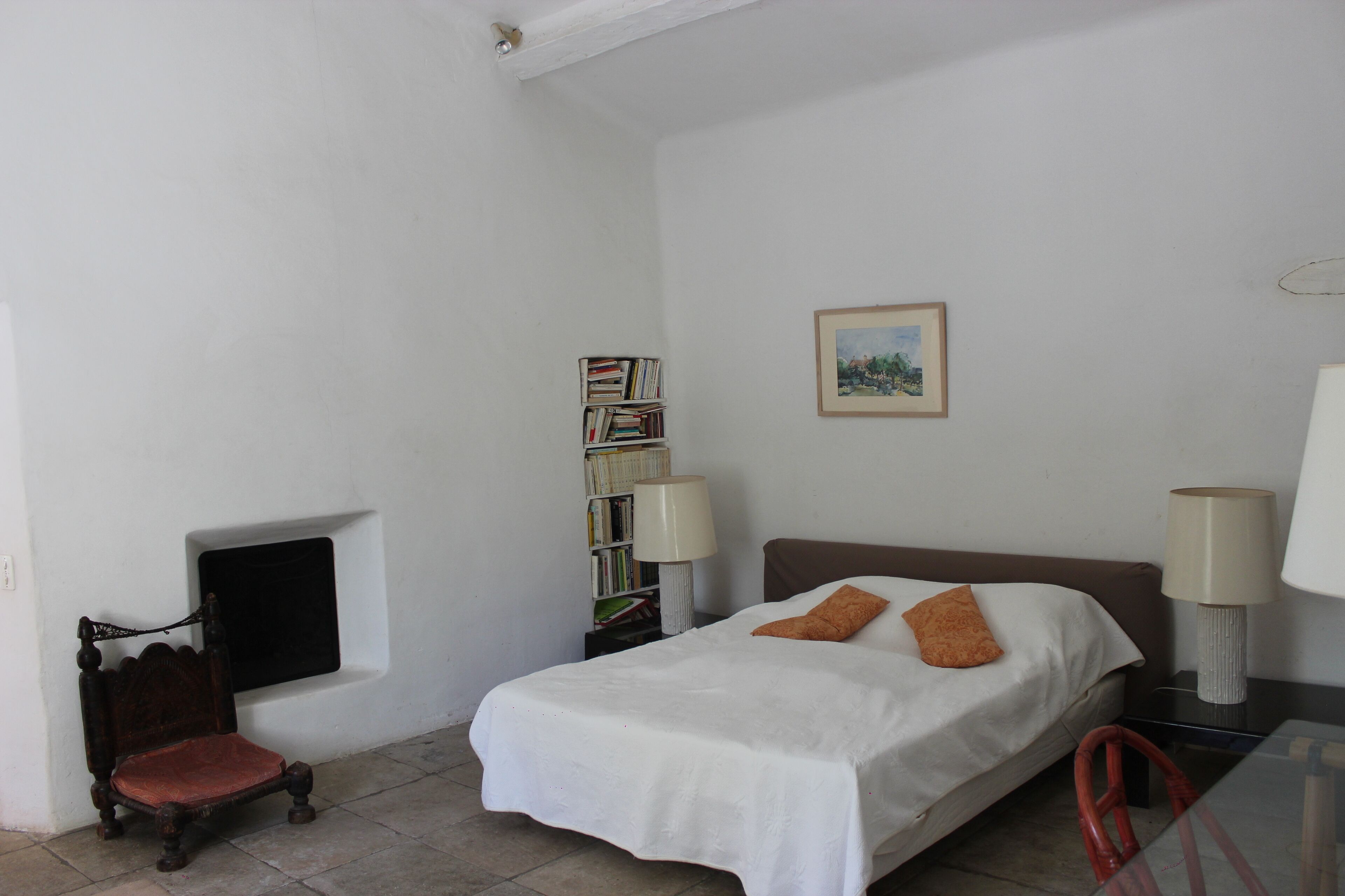 A bedroom with a single bed, a nightstand, a bedside table, a chair, a bookshelf, and a window.
