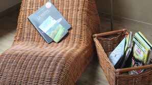 Un fauteuil de relaxation en osier, avec un coussin et un livre, un lampadaire et un panier rempli de magazines.