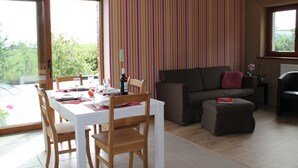 Un coin repas avec une table dressée pour deux personnes, un canapé et une fenêtre donnant sur un paysage verdoyant.