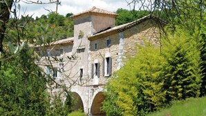 Un bâtiment en pierre, surmonté d’un toit en tuiles, entouré d’une végétation luxuriante et d’arbres.