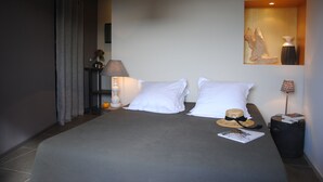 Un lit bien fait, avec des oreillers blancs, une couverture grise et un chapeau de paille posé sur le lit. Sur une table de chevet, on aperçoit une lampe, un vase rempli de fleurs et, au mur, un tableau encadré.