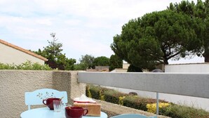 Une terrasse avec une table et des chaises, une petite table sur laquelle se trouvent un gâteau et des boissons, et un grand arbre en arrière-plan.