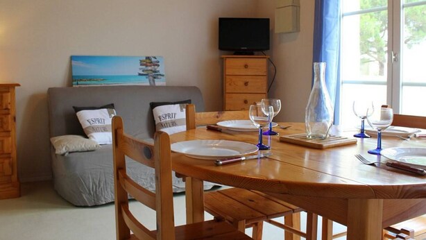Un coin repas avec une table en bois dressée pour quatre personnes, un canapé avec des coussins, un téléviseur posé sur un meuble et une fenêtre avec des rideaux.