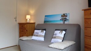 Une chambre à coucher avec un lit, deux oreillers, une table de chevet avec une lampe, une commode en bois et un mur orné d’un tableau représentant une plage et d’un panneau indicateur.