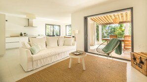 Ein modernes Wohnzimmer mit einem weißen Sofa, einem kleinen runden Tisch und einem Korbstuhl. Ein großes Schiebeglasfenster führt in einen Außenbereich mit einer Holzpergola und Blick auf Grünflächen.