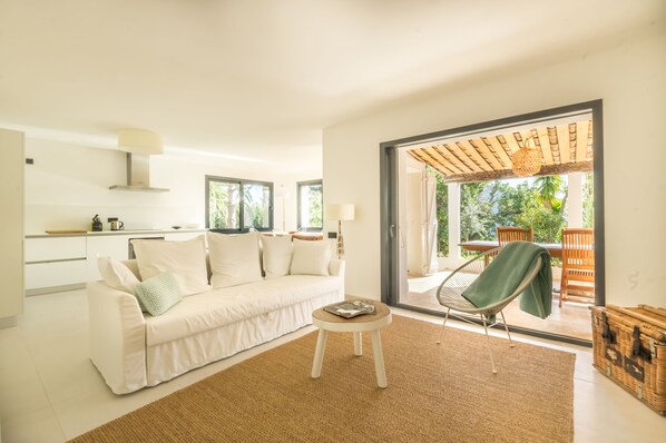 A modern living room with a white sofa, a small round table, and a wicker chair. There is a large sliding glass door leading to an outdoor area with a wooden pergola and a view of greenery.