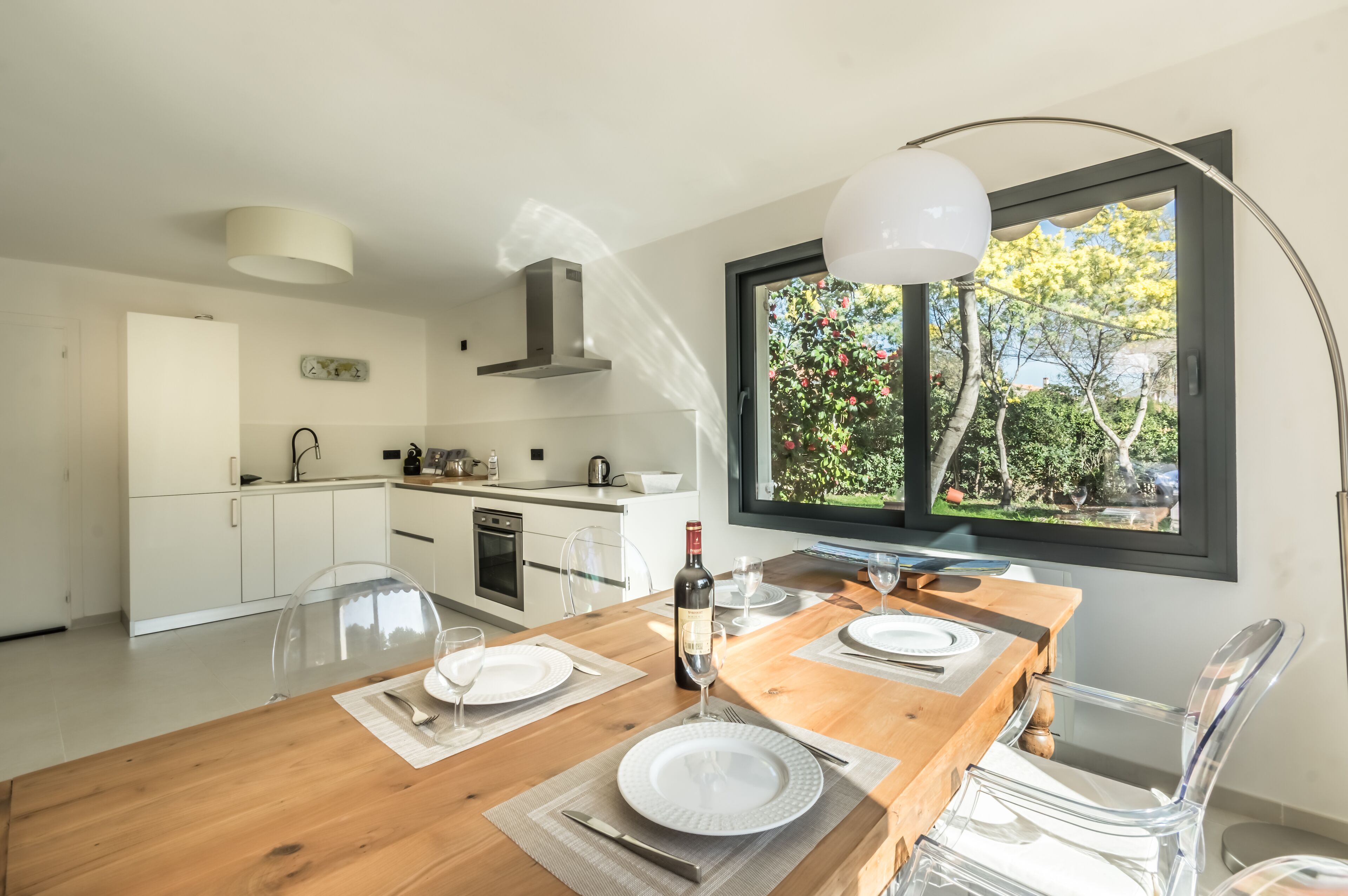 Une cuisine moderne avec des meubles blancs, une table à manger en bois pour deux personnes et une grande fenêtre donnant sur un paysage arboré.