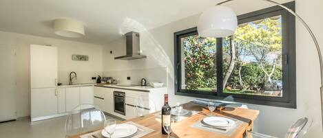 Une cuisine moderne avec des meubles blancs, une table à manger en bois pour deux personnes et une grande fenêtre donnant sur un paysage arboré.