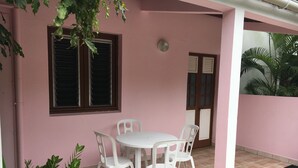 A pink exterior wall with a window and a door, a white round table with four chairs, and a potted plant. - APARTMENT F3 bottom of villa to rent for holidays (ANSES D'ARLET)