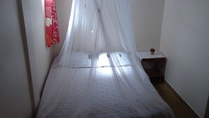 A small, narrow room with a bed covered by a sheer white canopy. A small wooden nightstand with a decorative item is visible to the right of the bed. - APARTMENT F3 bottom of villa to rent for holidays (ANSES D'ARLET)