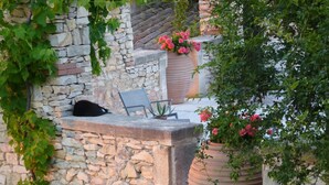 Una pared de piedra con un gato negro sentado en un alféizar, una silla y grandes macetas de terracota con flores.