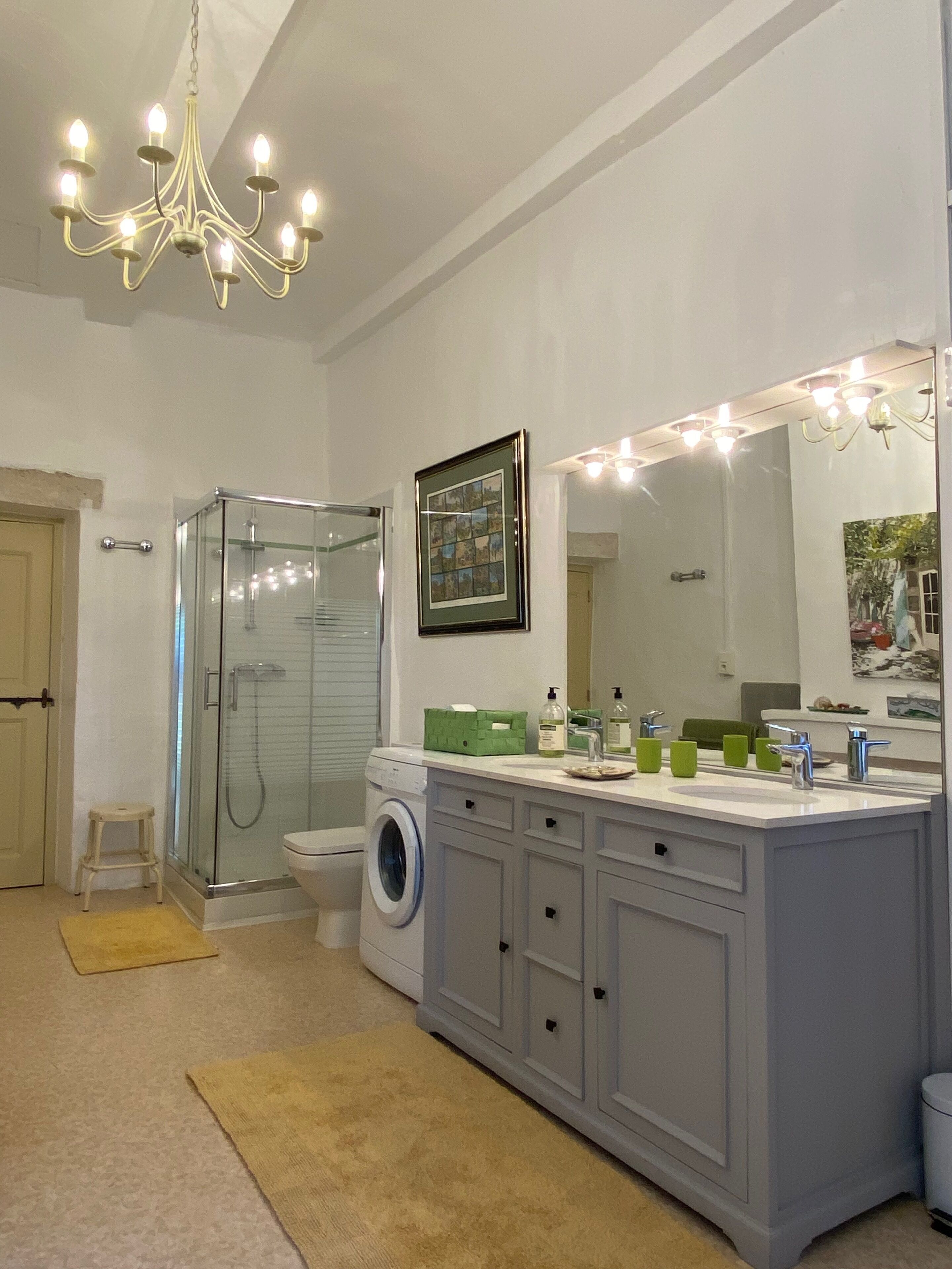 A modern bathroom with a glass shower enclosure, a built-in washing machine, a large vanity with a mirror, and a chandelier.