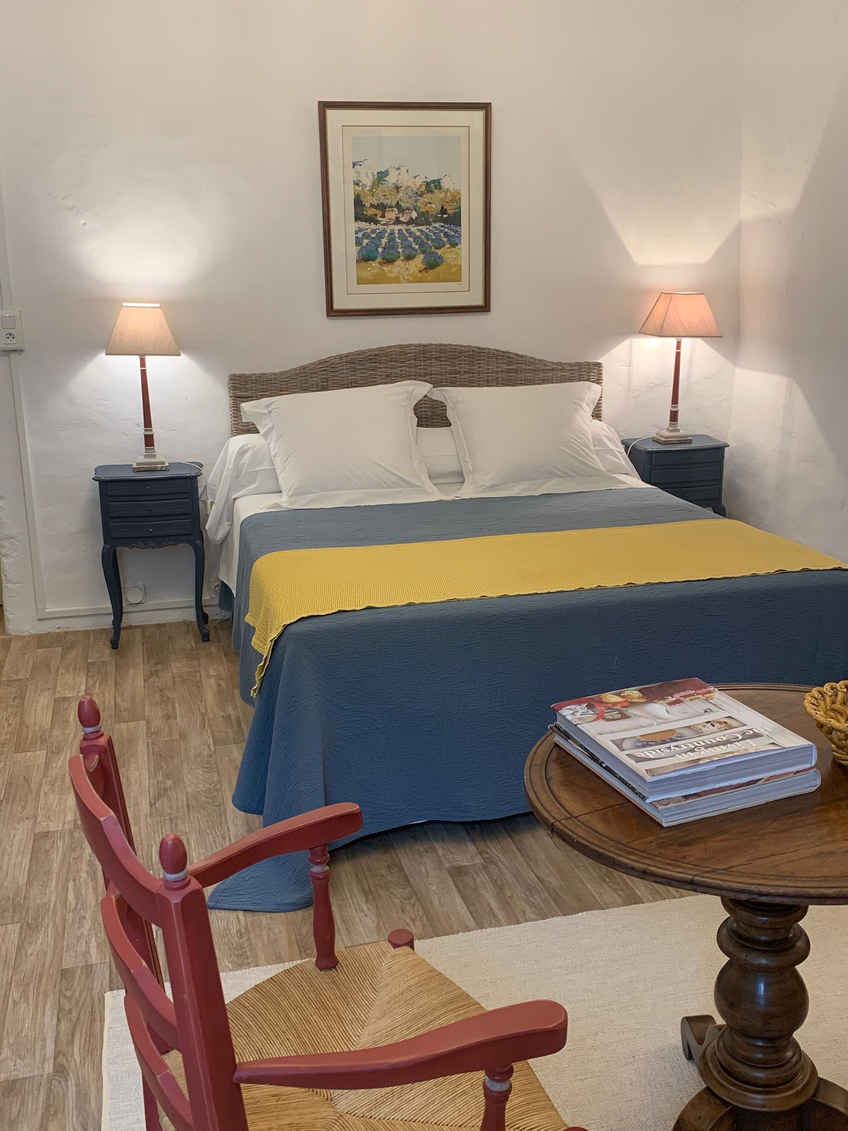 Un dormitorio ordenado con una cama, dos mesitas de noche, una silla, una mesa con revistas y un cuadro en la pared.