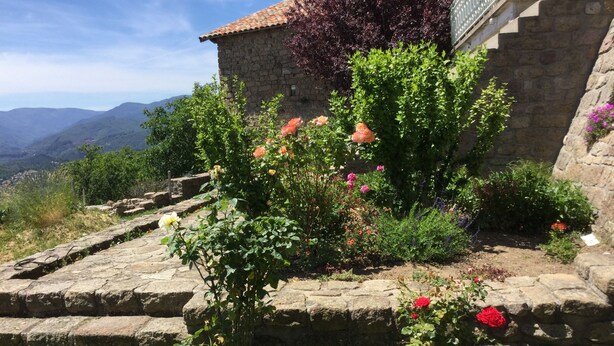 A stone building with a tiled roof, surrounded by a garden with various flowering plants and a stone pathway. - Quiet cottage with beautiful view (Chirols)
