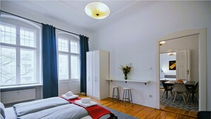 Ein Schlafzimmer mit Bett, einem Fenster mit Vorhängen, einem Kleiderschrank, einem Schreibtisch mit Stuhl und einem Essbereich mit Tisch und Stühlen.