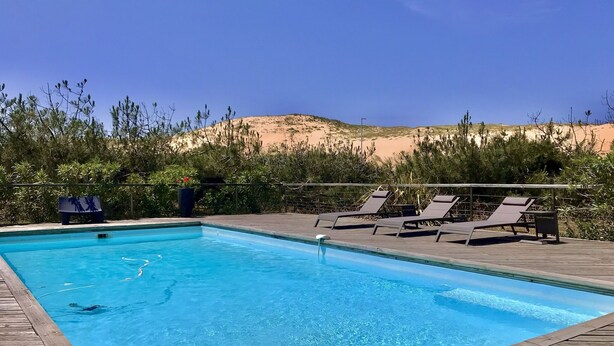 A rectangular swimming pool with clear blue water, surrounded by a wooden deck. There are lounge chairs and a sign with a silhouette of people. The background features a desert landscape with sparse vegetation and a clear sky. - Contemporary villa -Exceptional view of the dune-Private pool (Soorts-Hossegor)