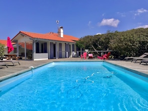 A rectangular swimming pool with clear blue water, surrounded by a wooden deck. There are lounge chairs and a red umbrella on the deck.