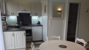Eine kompakte Küche mit weißen Schränken, einem eingebauten Backofen und einem Kühlschrank. Es gibt einen Esstisch mit Stühlen und einen kleinen Teppich.
