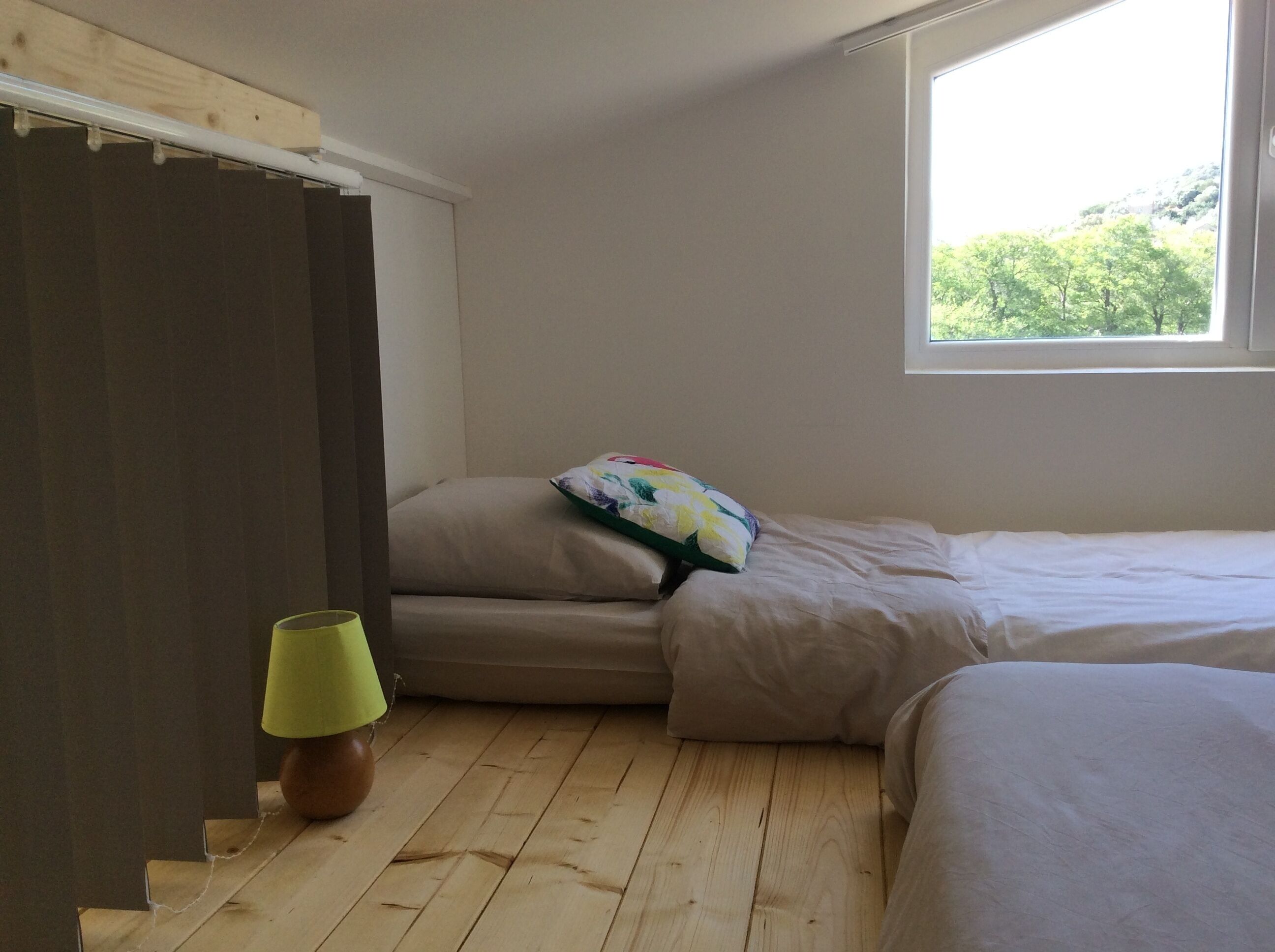 A bedroom with a wooden floor, a bed with white bedding, a window with a view of greenery, and a lamp on the floor.