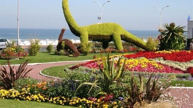 Un jardin aménagé avec une sculpture de dinosaure, une variété de fleurs aux couleurs vives et un sentier.