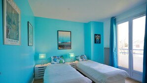 Une chambre avec deux lits, une table de chevet avec une lampe, un tableau encadré au mur et une fenêtre avec des rideaux.