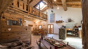 Ein rustikales Holzinterieur mit einem Essbereich, der einen langen Holztisch mit gedecktem Tisch für eine Mahlzeit, eine gemütliche Sitzecke mit einem Fellüberwurf und eine im Hintergrund sichtbare Küche zeigt.