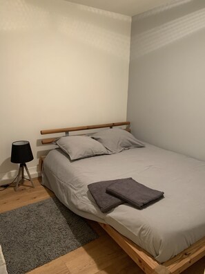 A single bed with a wooden frame, a grey blanket, and two pillows. There is a small table with a lamp beside the bed. - Small independent apartment in farmhouse with air conditioning (Chavanod)