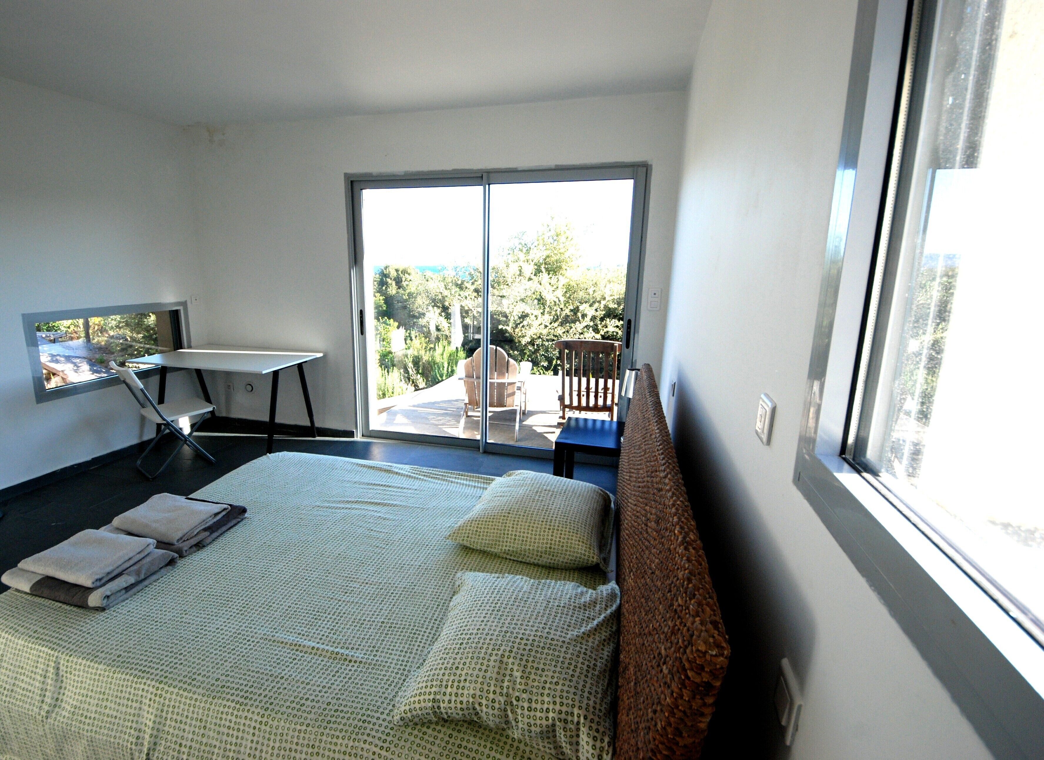 A bedroom with a bed, a desk, and a chair. There is a sliding glass door leading to an outdoor area with a table and chairs.