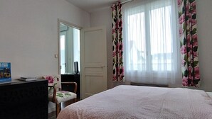 Ett hotellrum med en säng, ett skrivbord med en dator, en stol, en spegel och ett fönster med blommiga gardiner.