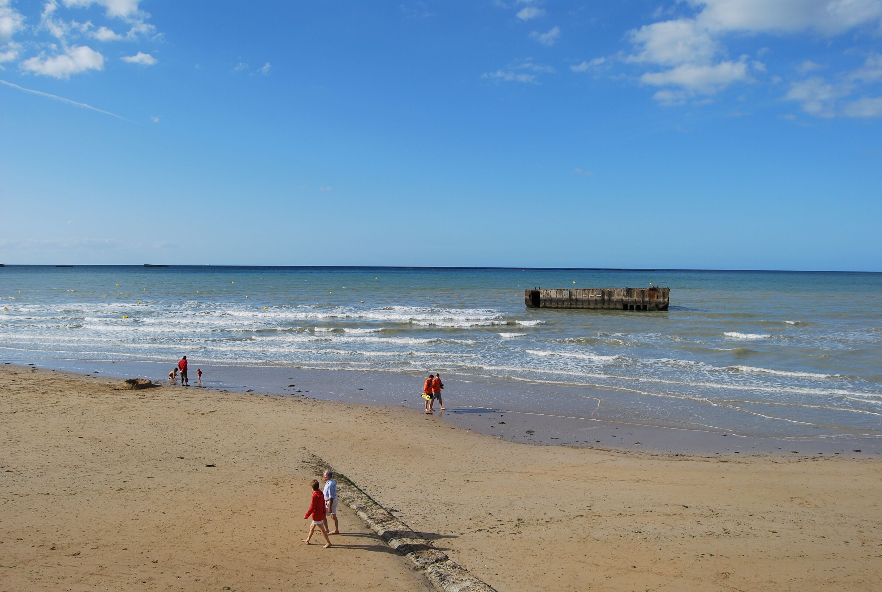 A beach with a wooden pier extending into the water, sandy shore, and people walking along the beach.